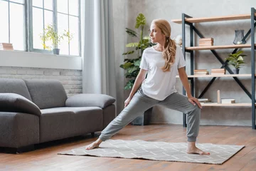 Foto op Plexiglas Home stretching, yoga exercises. Mature Caucasian woman in sporty clothes working out, doing physical training at home, losing weight, burning calories © InsideCreativeHouse