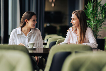 Fototapeta premium Two cheerful female friends are sitting in a coffee shop, chatting and having a coffee together.