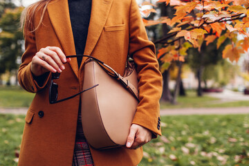 Close up of stylish woman holding handbag wearing orange blazer in autumn park. Fall female clothes...