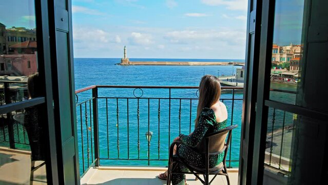 A beautiful girl in a dress enjoying the view from the hotel balcony in Chania, Crete. Young woman overlooking colourful Old Venetian Port of Chania surrounded by turquoise-blue water, mosque landmark
