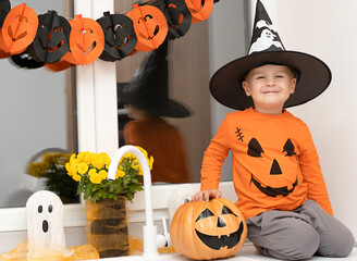 Halloween concept. A small, cheerful, handsome boy in a wizard's hat and an orange sweater sits on a table in the kitchen against the background of a pumpkin, a ghost and a bouquet of yellow flowers.