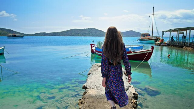 Beautiful girl enjoying the view of the blue crystal water with boats on the Mediterranean Sea. A woman walking on the coastal Greek island of Crete with turquoise water and ships on the seashore.