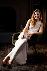 Obraz premium In the shadows, a young woman, her hair flowing, rests on a chair, casting a smile towards the camera lens in the darkness of the room