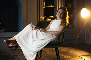 Surrounded by darkness, a young lady with flowing hair grins at the camera from her seat, creating a vivid contrast in the dimness of the room
