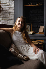 Obraz premium A young woman in a long white dress with long hair sits on the sofa smiling and looking at the camera. concept, have a good time