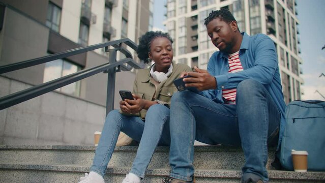 Two African American colleagues sitting on stairs and sharing social data. Showing beautiful woman his personal account in web. Talking about what they see on screens. Relaxing in middle of city.