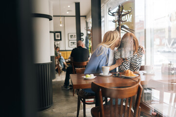 Wide shot of two best girlfriends sitting at indoors cafe by window and talking. Blonde female...