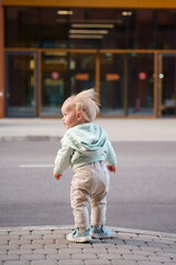 Cute blonde kid baby is standing by the road in the city, getting ready to cross the road, alone