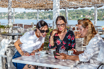 Fototapeta na wymiar Three cheerful girlfriends on a coffee break, talking on a restaurant terrace by the river. Woman showing pictures on her phone to her colleagues.