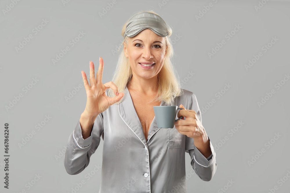 Poster Mature woman in pajamas with cup of coffee showing OK gesture on grey background