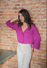 Beautiful young brunette woman in a purple blouse and white pants posing indoor against a brick wall.Portrait of a beautiful young brunette woman in a purple jacket 