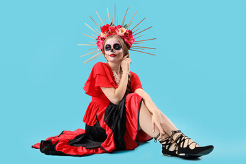 Young woman with painted skull on her face sitting against blue background. Celebration of Mexico's Day of the Dead (El Dia de Muertos)