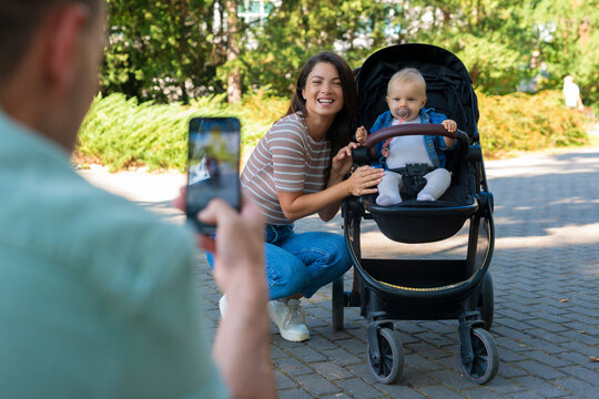 A husband is holding a phone and taking pictures of his wife and baby in the park