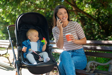 The baby is sitting in the stroller and holding a toy while mom is sitting on the bench and talking...