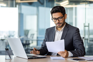 Portrait of serious pensive businessman behind paperwork, financier looking at documents, papers and contracts, thinking about solutions to set tasks, man inside office in business suit with laptop.