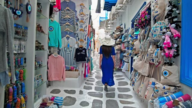 A tourist woman with a summer hat walking on the narrow streets with luxury shops in  Mykonos. Beautiful girl with a blue dress shopping for souvenirs, gifts and traditional Greek clothes in Mykonos.