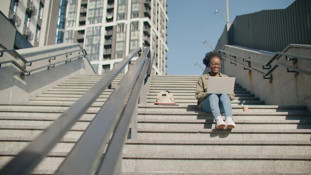 Busy African American girl with glasses using her laptop, typing and scrolling or browsing something on websites. Sitting on stairs in modern city in sunny day. Connection to Internet.