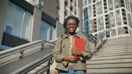 Happy African American woman with backpack standing on stairs and looking into camera. Positively smiling while holding red notebook or diary. Getting ready to start productive day.