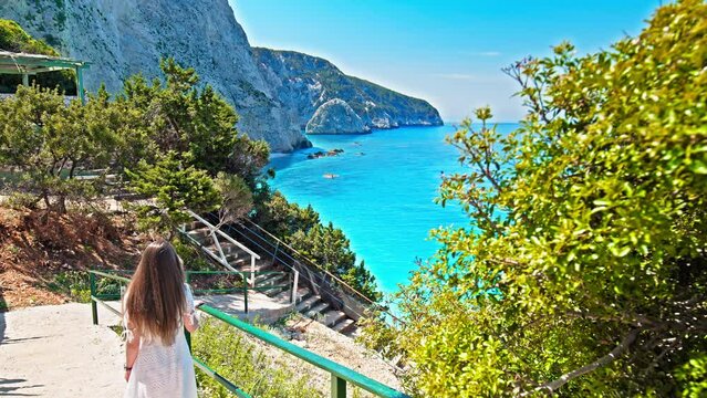 A beautiful woman in a white dress walks down the stairs to see Porto Katsiki beach. Female tourist relax at the narrow beach with picturesque views, turquoise water closely bordered by steep cliffs.