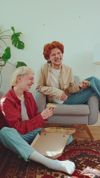 Laughing gay guys with dyed hair feeding each other pizza and talking at home 