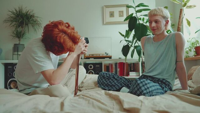 Pretty young gay couple with dyed hair taking pictures on camera on the bed at home