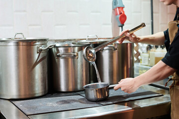 Hands of young male chef in apron pouring hot water into ladle while standing by his workplace in the kitchen of modern cafe or bar
