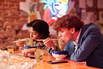 Young hungry man with curly hair bending over table with bowl while eating tasty ramen cooked by chef of Japanese cafe or restaurant