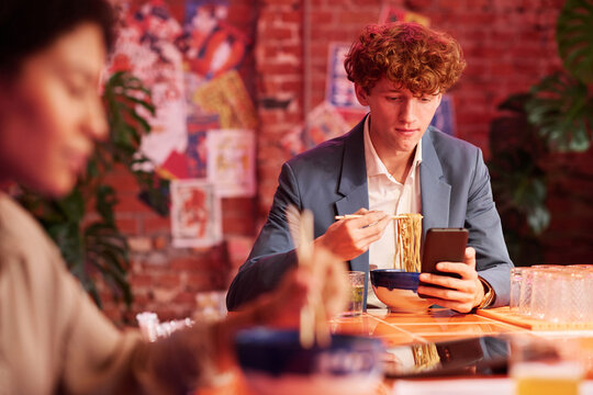 Focus on young curly man in white shirt and grey suit having ramen and watching online video in mobile phone while having lunch in cafe