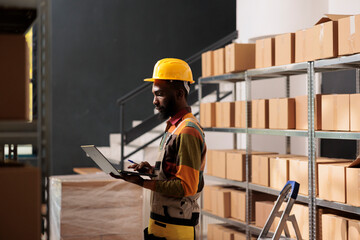 African american worker holding laptop computer, analyzing products checklist in warehouse. Storage room employee wearing protective overall while preparing customers orders before delivery goods