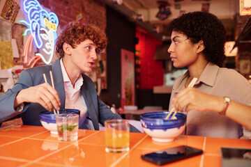 Two young intercultural colleagues or dates having chat at lunch break in Japanese cafe or restaurant while eating ramen and looking at one another
