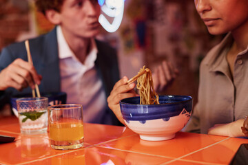 Focus on appetizing Japanese ramen on wooden chopsticks held by girl over bowl during lunch break with her colleague or boyfriend in cafe