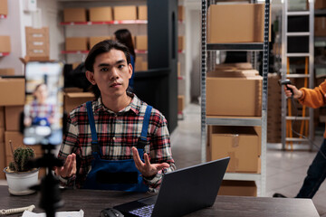 Postal service shipment manager filming order tracking operation on mobile phone camera. Warehouse asian employee sitting at desk in storage room and advertising goods for social media