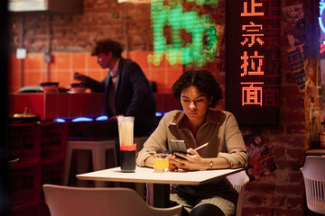 Serious girl texting in smartphone in cafe with hieroglyph meaning best ramen while sitting by...