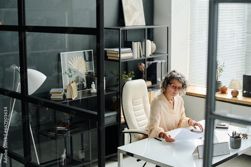Poster Mature female boss with financial paper sitting by workplace in office and looking through points of contract while checking data before entering