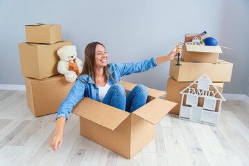Happy woman drinks champagne from a glass while sitting inside a cardboard box in new apartment.