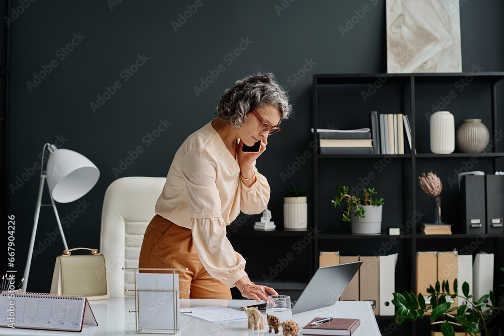 Aufkleber side view of busy mature female boss with smartphone by ear bending over desk and typing on laptop k