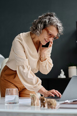 Mature elegant woman in eyeglasses pressing buttons of laptop keyboard while bending by desk and...