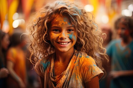 Portrait of happy smiling preteen girl with blond long curly hair at holi colors festival Face, orange tee shirt and hair painted with holi orange and blue colors. Colorful background.