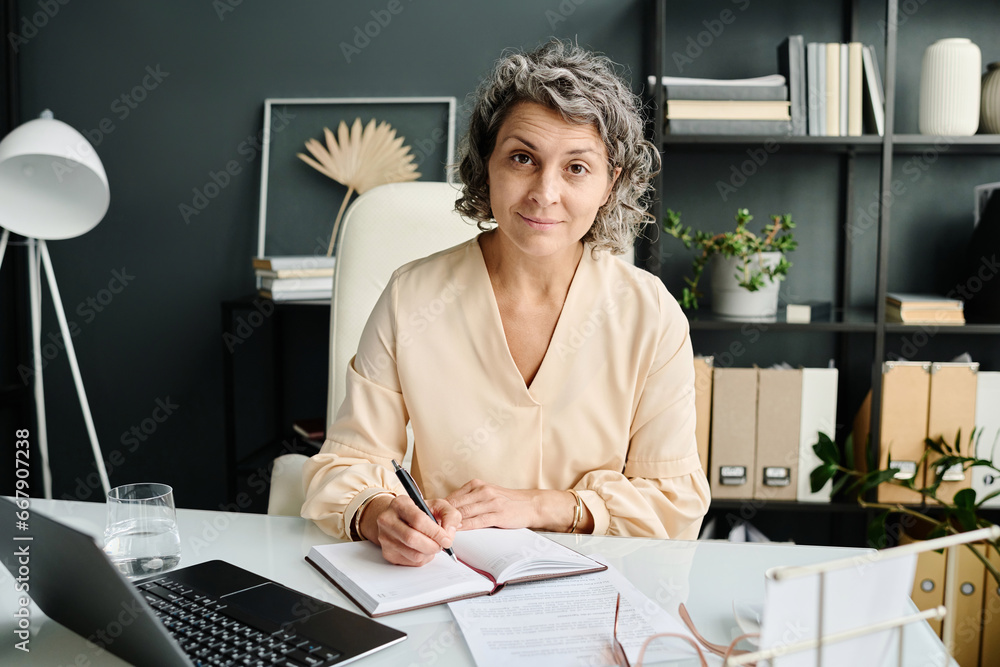 Poster experienced female director of business company looking at you with smile while sitting by her workp