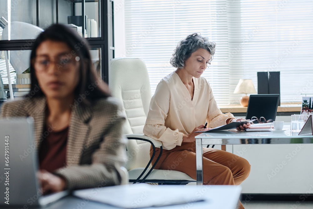 Poster Serious mature female director of company reading business document and checking its points while sitting by desks in front of subordinate