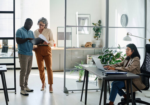 Mature businesswoman and African American male employee discussing document while standing in the center of modern coworking area