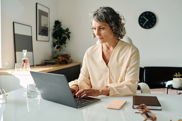 Serious mature female director of company looking at laptop screen and typing on keyboard while...