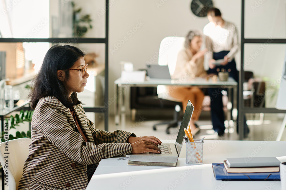 Poster side view of young female analyst looking at online data on laptop screen and typing on keyboard whi