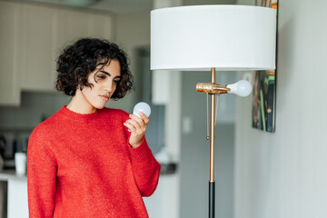 Curly middle eastern woman holding light bulb for chandelier and looking on it at home