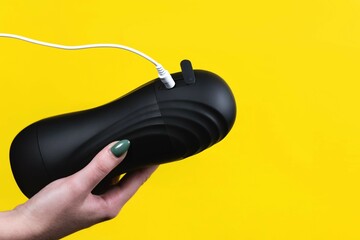 Sex toys for men. Close-up of a massager for male masturbation with a connected charging cable in female hands on a yellow background. Sex shop Adult store - obrazy, fototapety, plakaty