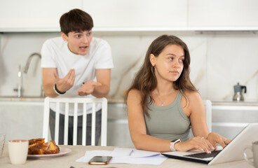 Annoyed young girl doing business on notebook while guy is brawling with her at the kitchen table
