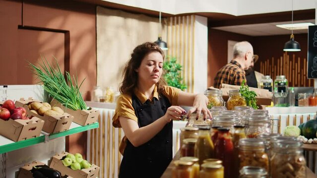 Portrait of smiling woman working in zero waste supermarket, adding pantry staples on shelves. Cheerful merchant restocks local grocery store with organic food in reusable jars, close up