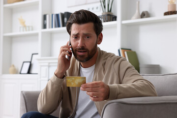 Fototapeta premium Upset man with credit card talking on phone in armchair at home. Be careful - fraud