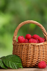 Wicker basket with tasty ripe raspberries and green leaves on table against blurred background, closeup