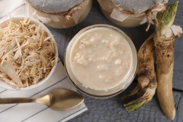 Spicy horseradish sauce in jars, roots and spoon on grey wooden table, flat lay